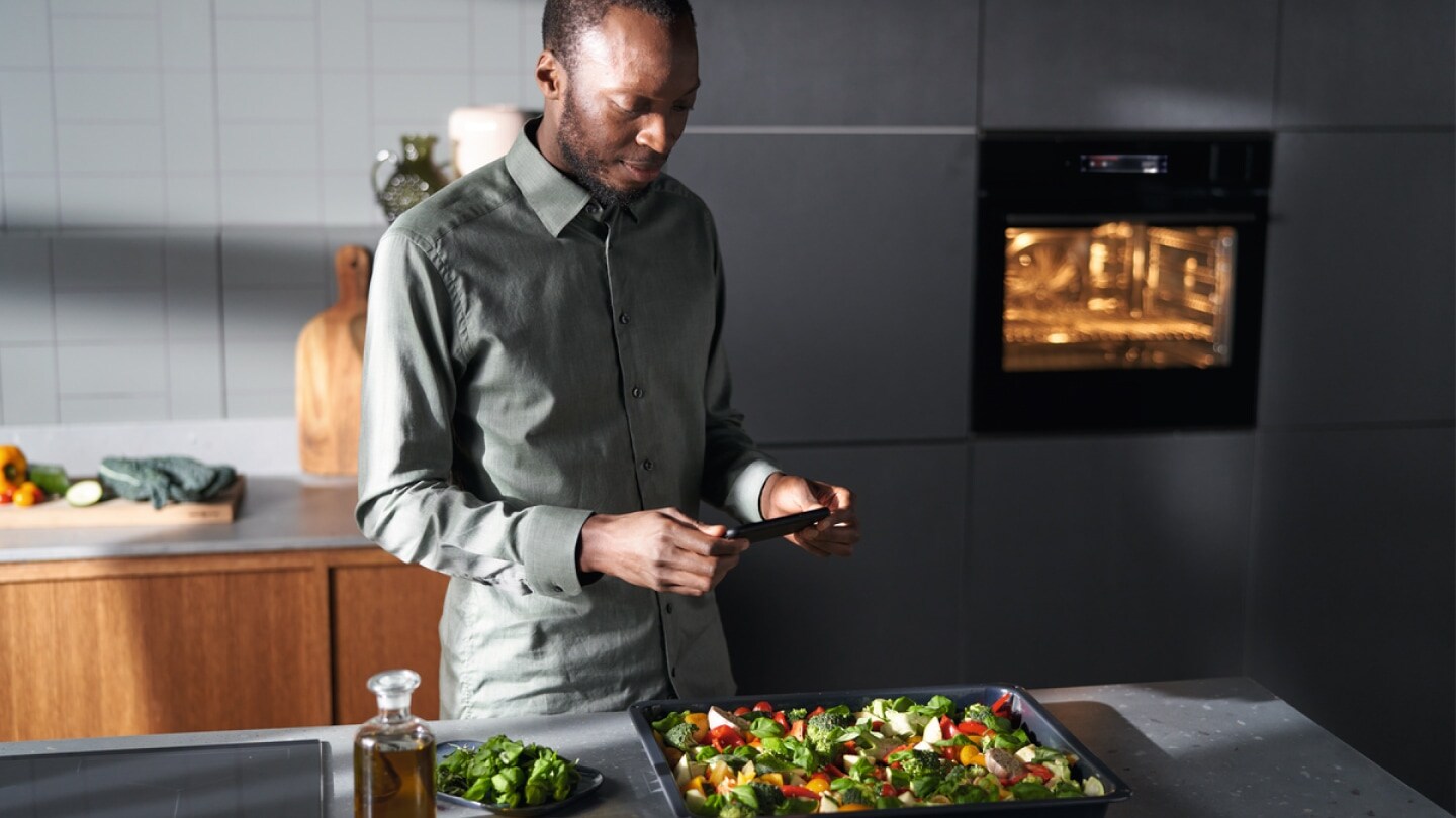 A man looking at his phone and cooking vegetables on a hob